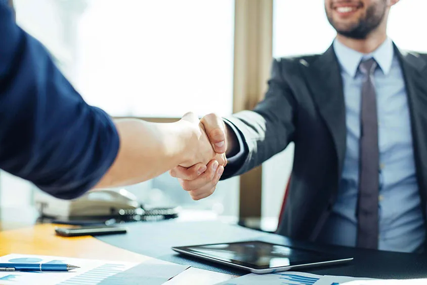 Buyer and supplier shaking hands across a desk covered with a tablet, contracts and cost charts