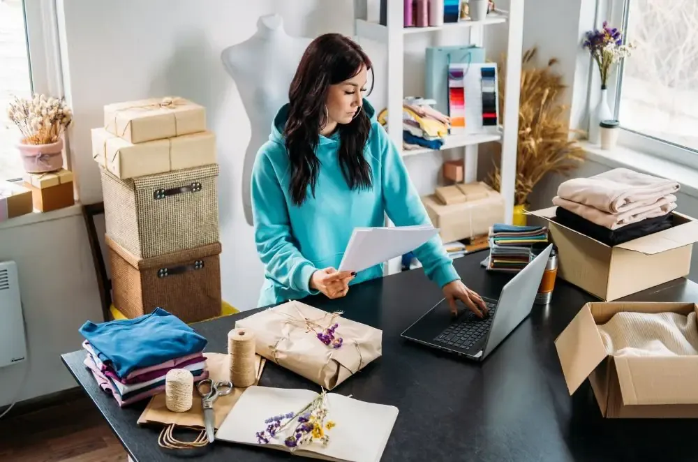 E-commerce founder reviewing a supplier document beside a laptop, packed parcels, folded textiles and colour swatches
