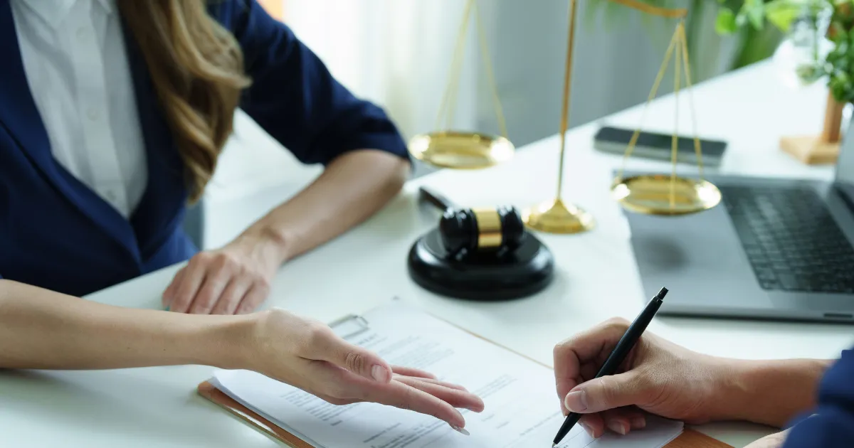 Hands signing a compliance document on a clipboard with a gavel and scales of justice on the desk
