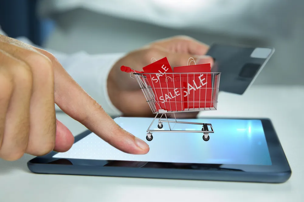 A shopper pointing at an online store on a tablet, with a mini shopping cart of sale items